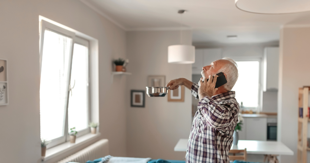 Homeowner catching water leaking from ceiling while calling a plumber, illustrating the urgency for professional leak detection during an active plumbing failure.